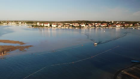 Aerial-panorama-of-a-Nin-town-with-lagoon-and-following-boat-approaching-sandy-beach-in-sunset