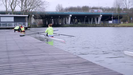 Rowers-preparing-to-starting-training-and-pushing-off-the-jetty-ready-to-start,-with-a-view-down-the-canal-in-Utrecht,-Netherlands-and-a-road-bridge-in-the-background