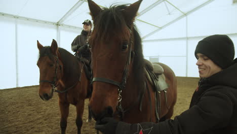 Trainer-shows-teeth-of-brown-horse-adjusting-bridle-by-second-man-on-horseback