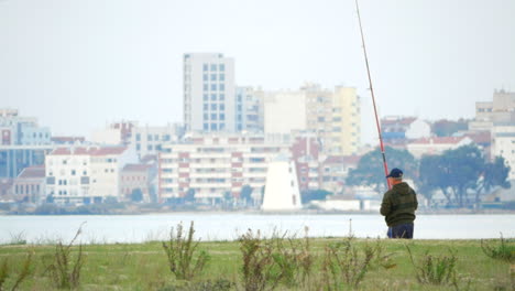 Old-Fisherman-On-Riverside-Reeling-Fishing-Rod-Wity-City-Buildings-In-Background