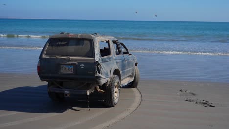 Slow-Motion-Shot,-Van-Parked-on-the-shoreline-of-San-Juanico,-Baja-California-Sur,-Mexico,-on-a-bright-sunny-day-scenic-view-of-the-blue-sea-and-birds-flying-in-the-background