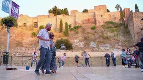 Chico-Con-Máscara-Jugando-Burbujas-Con-Niños-Frente-Al-Antiguo-Teatro-Romano-En-Málaga,-España---Nivel-Del-Suelo,-Tiro-Estático