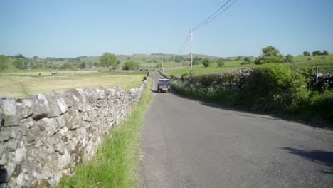 Static-slow-mo-shot-of-old-blue-Land-Rover-defender-driving-through-British-country-lanes-featuring-iconic-drystone-walls-on-a-summers-day-with-bright-blue-clear-skies