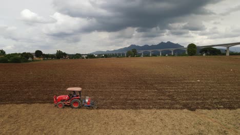 Luftaufnahmen-Eines-Traktors,-Der-Ein-Ackerland-Pflügt-Und-Nach-Links-Geht,-Wobei-An-Einem-Bewölkten-Tag-Auch-Eine-Hochgeschwindigkeitsstrecke-Und-Berge-Am-Horizont-Sichtbar-Sind