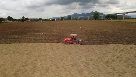 Deslizándose-Hacia-La-Izquierda-Imágenes-Aéreas-De-Un-Agricultor-Que-Trabaja-Duro-Conduciendo-Un-Tractor-Rojo-A-Través-De-Tierras-De-Cultivo,-Labrando-Y-Preparándose-Para-Un-Nuevo-Lote-De-Plantas,-Red-Ferroviaria-De-Alta-Velocidad-En-El-Horizonte