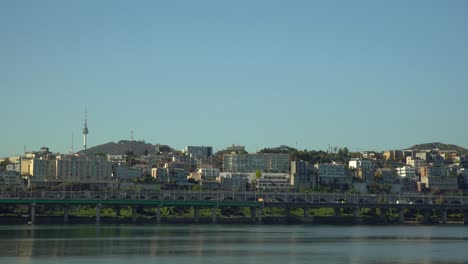 N-Seoul-Namsan-Tower-over-Yongsan-gu-district-urban-skyline,-Daytime-traffic-on-Gangbyeonbuk-ro-expressway-by-Han-river---wide-static-copy-space