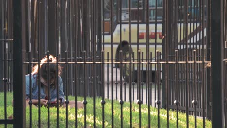 Junges-Mädchen-Mit-Maske-Und-Telefon-Im-Park,-Umgeben-Von-Einem-Zaun