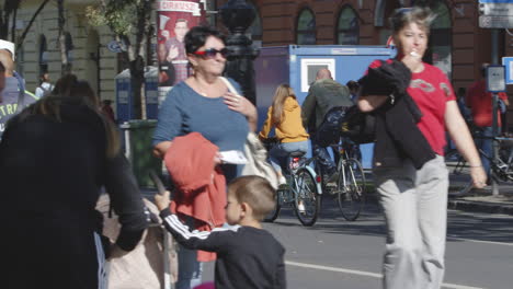 A-couple-ride-their-bicycles-away-on-busy-city-street-with-pedestrians-in-Budapest,-Hungary