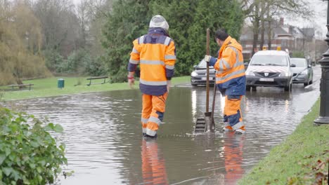 A-Municipal-worker-signals-a-car-to-slow-down-through-a-flooded-residential-street,-unplugging-drain