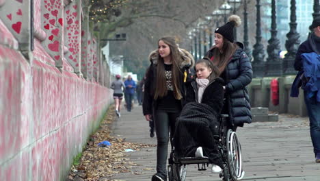 A-slow-motion-shot-of-a-woman-in-a-wheelchair-accompanied-by-two-others-look-at-the-thousands-of-red-painted-hearts-with-messages-to-lost-loved-ones-at-The-National-Covid-Memorial-Wall