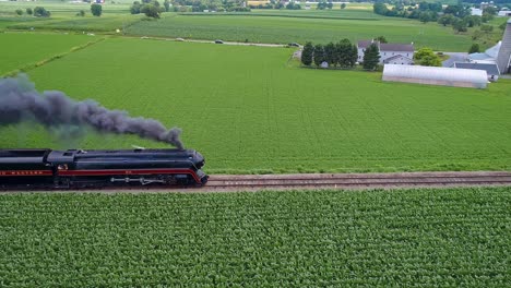 Una-Vista-Aérea-Paralela-De-Un-Antiguo-Tren-De-Pasajeros-De-Vapor-Que-Viaja-Con-Humo-Negro-A-Través-De-Tierras-De-Cultivo-En-Un-Día-Soleado