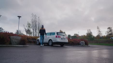 Joven-Alto-Con-Gorra-Entra-En-Su-Auto-Blanco-En-Un-Pequeño-Estacionamiento,-Plano-Estático