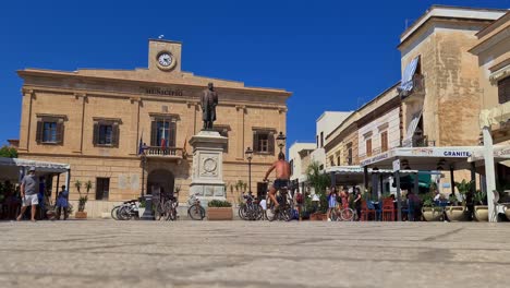 ángulo-Bajo-Y-Cámara-Lenta-Del-Hombre-En-Bicicleta-En-La-Plaza-Piazza-Europa-En-La-Isla-Favignana-Con-El-Ayuntamiento-En-El-Fondo,-Sicilia