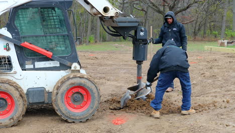 Tres-Trabajadores-De-La-Construcción-Que-Trabajan-Con-Palas-Y-Sinfín-Hidráulico-Montados-En-Un-Cargador-De-Dirección-Deslizante-Para-Cavar-Un-Agujero-De-Poste-Para-El-Marco-De-La-Construcción-De-Un-Nuevo-Granero