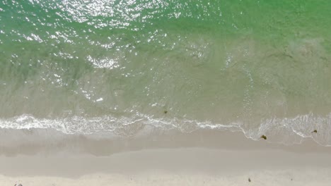 Low-altitude-birds-eye-view-over-waves-crashing-on-a-sandy-beach-during-a-bright-and-sunny-afternoon