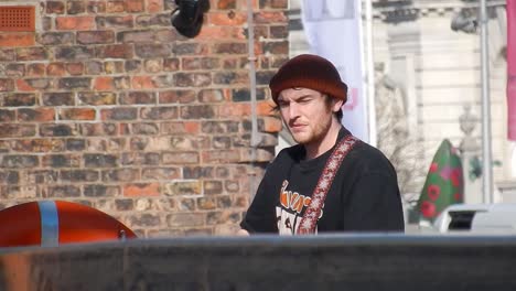 Young-male-busking-on-city-streets-with-guitar,-singing---wearing-hat