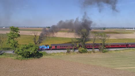 Vista-Aérea-De-Thomas-El-Tren-Echando-Humo-Negro-Y-Tirando-De-Coches-De-Pasajeros-Llenos-De-Niños-Felices-En-Un-Día-Soleado-Visto-Por-Un-Dron