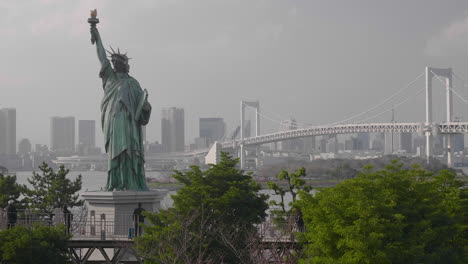 Los-Turistas-Caminan-Por-El-Paseo-Marítimo-De-Odaiba,-Con-La-Estatua-De-La-Libertad-Y-El-Puente-Del-Arco-Iris-Detrás-De-Ellos