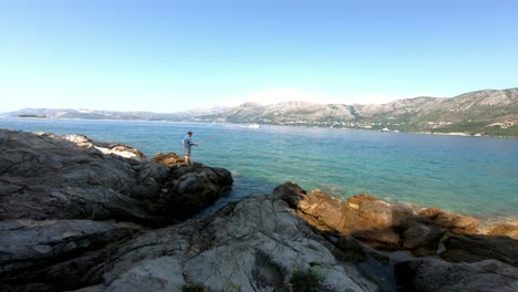 A-man-stands-and-fishes-on-the-rocky-coastline-of-the-Adriatic-while-powerboat-speeds-near-by