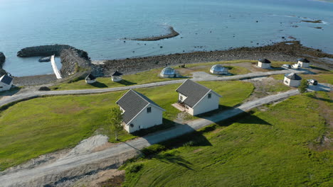 Aerial-view-of-houses-and-aurora-hut,-glass-igloos,-at-a-resort,-on-the-coast-of-the-Arctic-sea,-sunny-day,-in-Spaakenes,-Troms,-Nordland,-North-Norway---orbit,-drone-shot