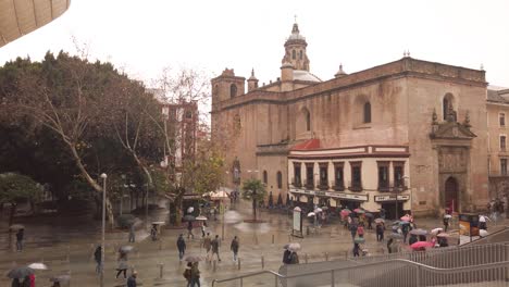 Cámara-Lenta,-La-Gente-Pasa-Por-La-Iglesia-En-La-Lluviosa-Sevilla,-Vista-Desde-Un-Punto-De-Vista