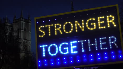 Un-Avión-De-Pasajeros-Pasa-Detrás-De-Un-Cartel-Iluminado-Que-Dice-&quot;más-Fuertes-Juntos&quot;-Y-La-Abadía-De-Westminster-Se-Ve-Al-Fondo-Por-La-Noche