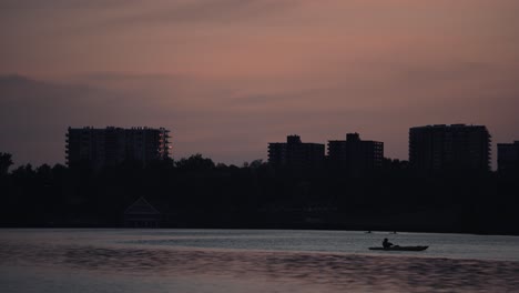 Silueta-De-Personas-En-Bote-En-El-Lago-De-Las-Naciones-Con-Edificios-En-El-Fondo-En-Sherbrooke,-Quebec,-Canadá-Al-Atardecer---Plano-General