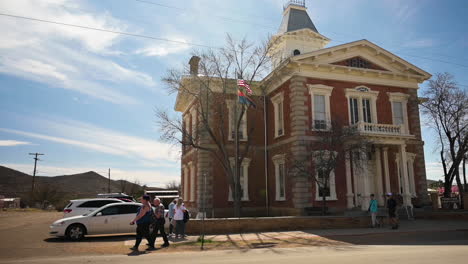 Menschen,-Die-Vor-Dem-Historischen-Cochise-County-Courthouse-Im-Tombstone-State-Park-In-Arizona,-USA,-Mit-Draußen-Geparkten-Autos-Und-Unter-Dem-Himmel-Wehender-Amerikanischer-Flagge-Spazieren-–-Schwenkaufnahme