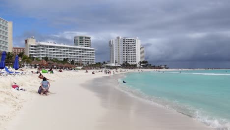 Establishing-shot,-people-relaxing-on-the-shore-and-swimming-on-the-beach-in-Yucatan,-Mexico,-Buildings,-wooden-huts-and-cloudy-sky-in-the-background