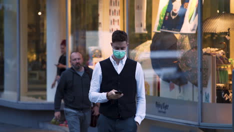 Ein-Eleganter-Junger-Mann-Mit-Grüner-Covid-19-Schutzmaske,-Schwarzer-Weste,-Weißem-Hemd-Und-Jeans-überquert-Die-Straße-Und-Ruft,-Geht-Auf-Die-Kamera-Zu,-Im-Hintergrund-Ein-Geschäft,-4k