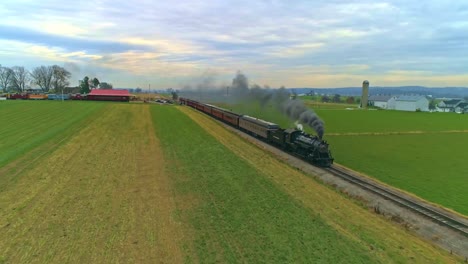 Una-Vista-Aérea-De-Un-Antiguo-Tren-De-Vapor-Restaurado-Y-Vagones-De-Pasajeros-Que-Se-Acercan-Con-Humo-Y-Vapor-A-Lo-Largo-De-Verdes-Tierras-De-Cultivo-Y-Campos-Vistos-Por-Un-Dron