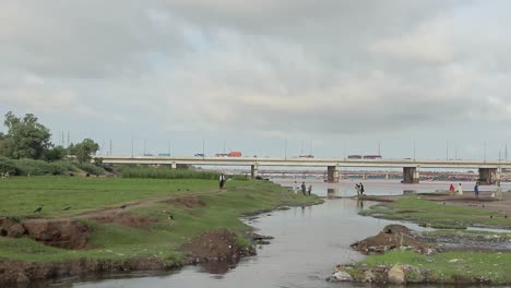 Gente-Caminando-A-Orillas-Del-Río-Ravi,-Vehículos-Viajan-En-El-Paso-Elevado-Del-Puente-En-Pakistán