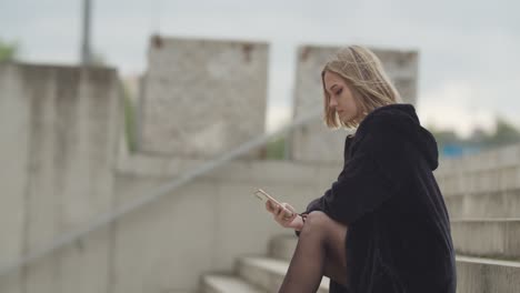 Attraktives-Junges-Blondes-Mädchen,-Das-An-Windigen-Tagen-Draußen-Auf-Der-Treppe-Sitzt-Und-Smartphone-Schaut