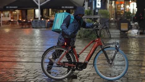 Deliveroo-Food-Fahrer-Mit-Thermorucksack-Und-Winteruniform-Steigt-Auf-Sein-Fahrrad-Und-Fährt-Auf-Der-Straße-Im-Stadtzentrum,-Um-Bestellungen-Für-Kunden-Und-Kunden-Auszuliefern