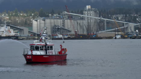 Bote-De-Bomberos-Canadiense-Rociando-Con-Agua,-Ejercicio-De-Entrenamiento-En-El-Río-Con-Fábrica-Industrial-En-Segundo-Plano