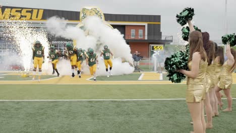 Slow-motion-college-football-players-break-through-paper-pregame-and-run-onto-the-field-as-champions-while-marching-band-and-cheerleaders-cheer-Stock-Video-Footage