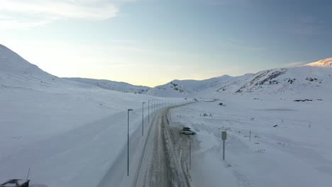 Camión-Del-Transporte-Tsl-Cruzando-La-Montaña-Hemsedal-En-Una-Carretera-Cubierta-De-Nieve-Al-Atardecer---Vuelo-Aéreo-Sobre-La-Carretera-Con-Un-Camión-Superando-A-Un-Dron-Y-Revelándose-En-Medio-Del-Clip