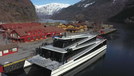 Catamarán-De-Pasajeros-Completamente-Eléctrico-Futuro-De-Los-Fiordos-Junto-A-Flam-Noruega---Antena-Acercándose-Lentamente-Y-Volando-Sobre-El-Barco-Con-El-Pueblo-De-Flam-Y-El-Fondo-De-La-Montaña