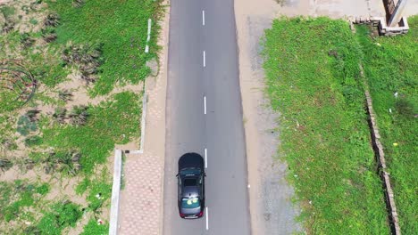 Imágenes-Aéreas-Que-Siguen-A-Un-Automóvil-Oscuro-De-Lujo-Con-Un-Automóvil-Blanco-Que-Lo-Sigue-En-Un-Camino-De-Concreto-Vacío,-Vegetación-Y-Arena-En-Ambos-Lados,-Viaje-Por-Carretera,-Al-Aire-Libre,-Techo-Solar,-Cocoteros,-Edificio,-Brillante