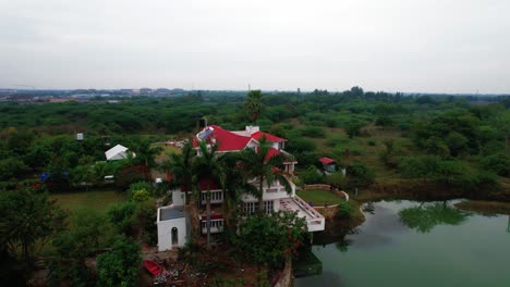 Vista-Aérea-De-360-Grados-De-Una-Casa-De-Vacaciones-Rodeada-Por-Un-Lago-Celestial,-Estacionamiento-Y-Privacidad,-Con-Lago-Y-Naturaleza-Verde-Encantada-En-Vadodara,-India