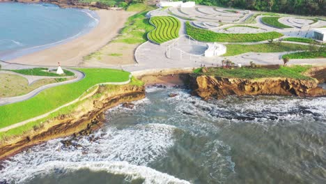 Tiro-Que-Se-Aleja-Lentamente-De-Una-Estructura-Expansiva-Justo-Al-Lado-Del-Mar,-Suaves-Olas-Blancas-Espumosas,-Arquitectura-Moderna,-Parches-Verdes-Brillantes,-Día-Soleado,-Acantilado-Visible,-Paisaje-Natural-Colorido