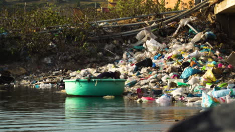 Hombre-En-Un-Bote-Vietnamita-Redondo-Inspeccionando-La-Orilla-Del-Río-Contaminada-Con-Basura-Plástica