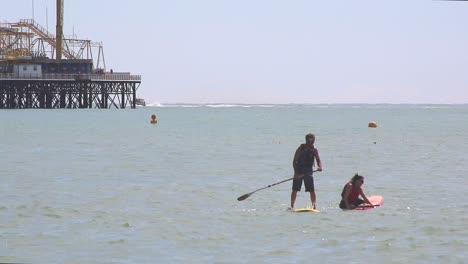 Brighton,-England---11.-August-2018:-Ein-Sportliches-Paar,-Das-An-Einem-Sonnigen-Tag-Vor-Den-Ausgebrannten-Skelettresten-Des-Hauptpiers-In-Brighton-Paddleboards-Zur-Erholung-Nutzt