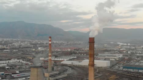 Rauchende-Industrieschornsteine,-Gebäude-Und-Berge-Als-Hintergrund---Luftaufnahme