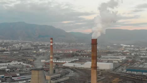 Industrial-chimney-smoking,-buildings-and-mountains-as-background---aerial-shot