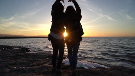 Hermanas-Haciendo-Un-Corazón-Juntas-Mientras-Disfrutan-De-Una-Hermosa-Puesta-De-Sol-Que-Cae-Bajo-El-Horizonte-De-Un-Estanque-De-Sal-Gigante