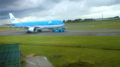 Avión-De-La-Aerolínea-Klm-Tirado-Por-Un-Camión-Remolcador-En-Una-Pista-Con-Nubes-Lluviosas-En-El-Fondo