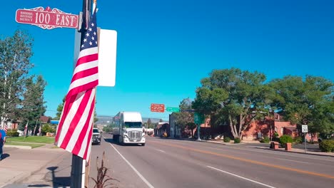 18-Wheeler-Fährt-Am-4.-Juli-In-Der-Stadt-Panguitch,-Utah,-Vorbei,-Mit-US-Flagge-Im-Vordergrund