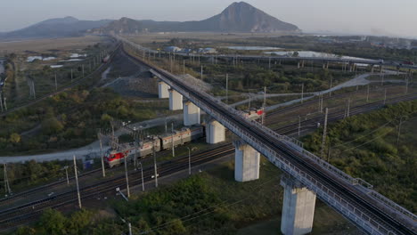 Russian-railway-train-locomotives-passing-under-the-car-bridge-with-mountains-in-the-background-on-the-sunset,-Russian-Federation