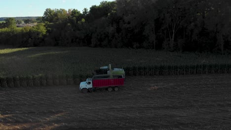 4k-Luftaufnahme-Mit-Schwenk-Nach-Links-Von-Der-Erntemaschine,-Die-Mais-In-Einen-Lastwagen-Häckselt,-Der-Im-Schatten-Zur-Goldenen-Stunde-Startet-Und-Sich-In-Die-Sonne-Bewegt